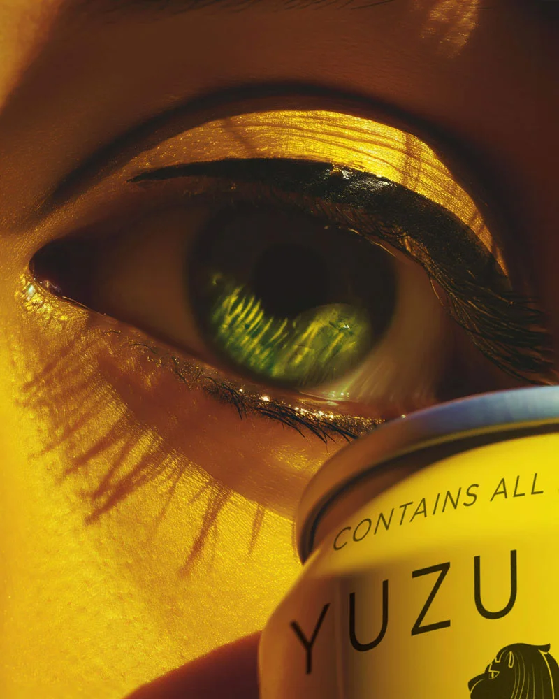 Close-up of a green eye with gold eyeshadow and black eyeliner, holding a yellow can labeled YUZU.